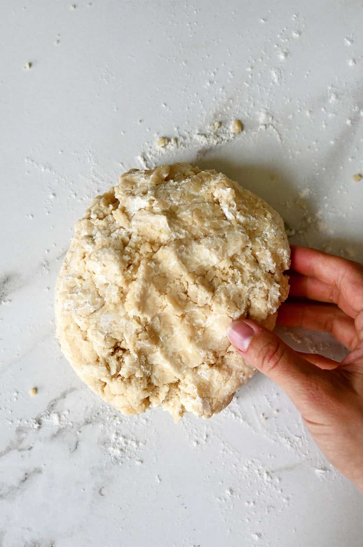A hand holds shaggy pie dough on a flour-dusted marble surface.