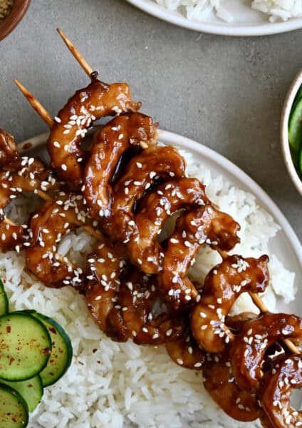 Honey garlic shrimp skewers on a plate containing steamed white rice and cucumber salad.