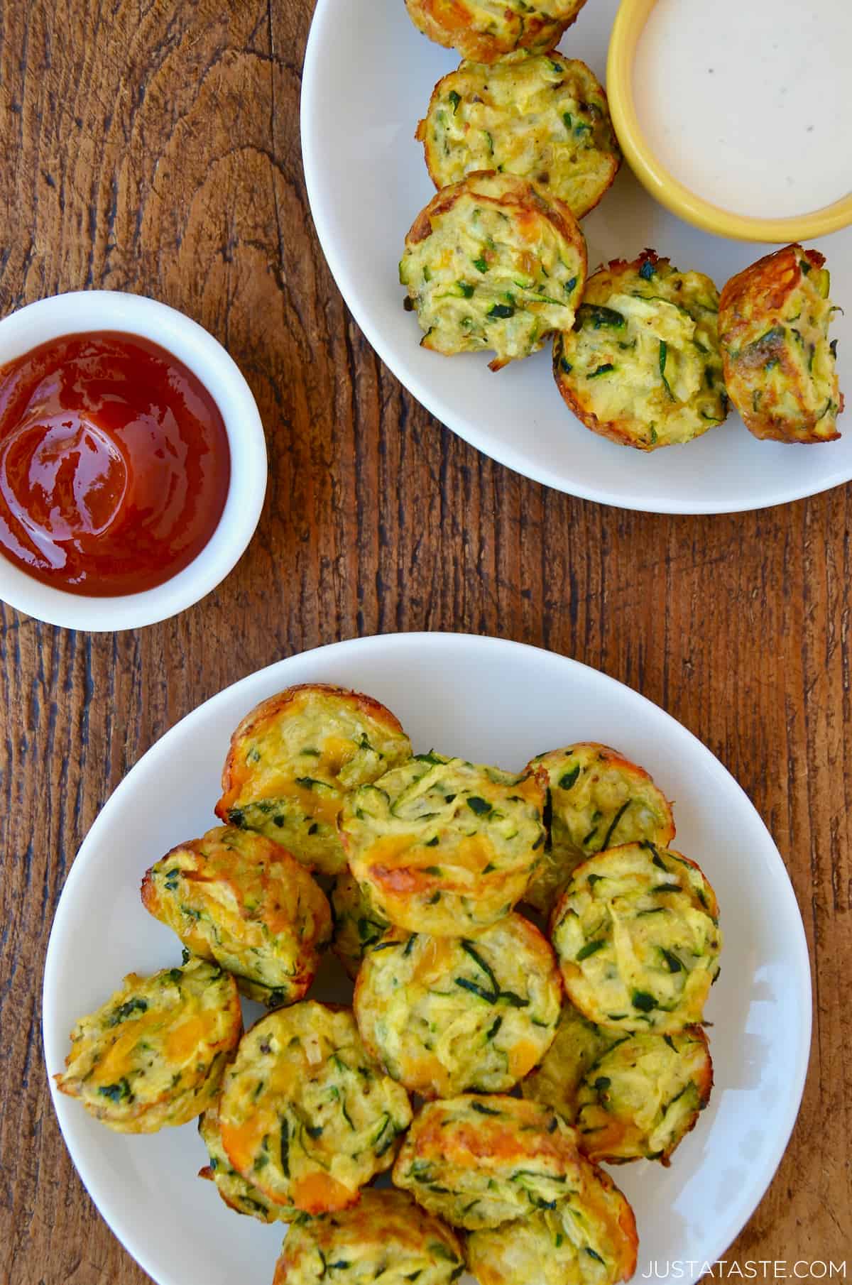 Two white dinner plates piled high with cheddar cheese zucchini tots next to a small bowl filled with ketchup and a small bowl containing ranch dressing.