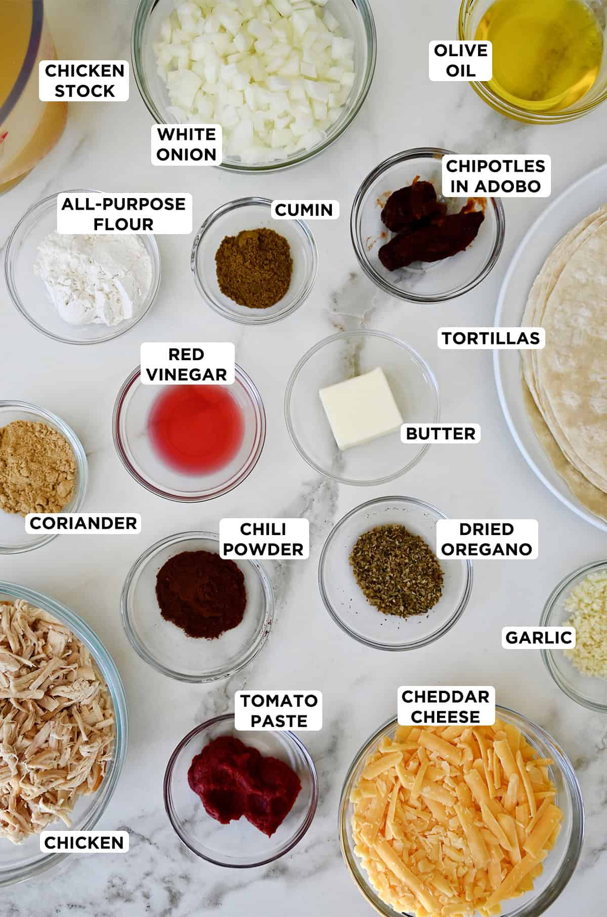 Various sizes of glass bowls containing ingredients to make rotisserie chicken enchiladas, including butter, flour, chicken stock, chipotles in adobo, tomato pasta, spices, red wine vinegar, onion, minced garlic, rotisserie chicken, shredded cheese and tortillas.