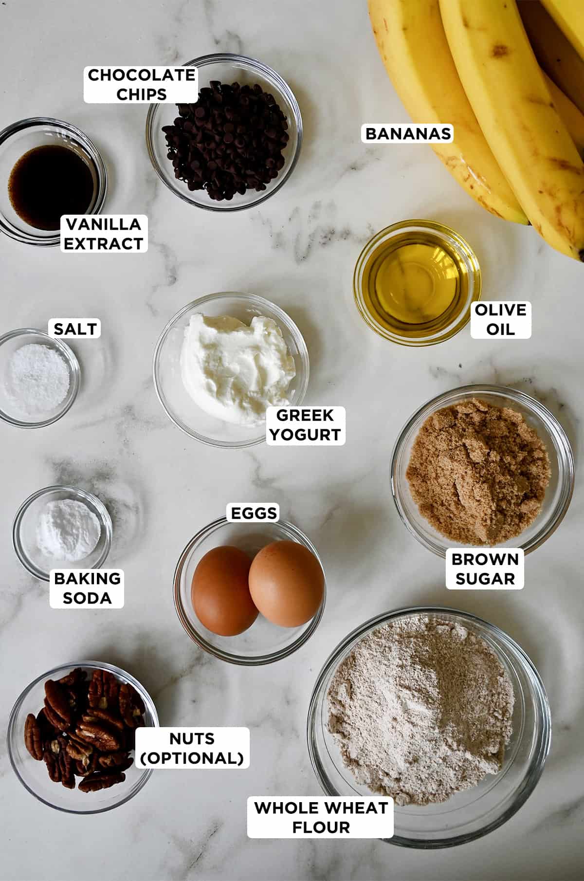 Glass bowls containing the ingredients needed to make whole wheat banana bread, including whole wheat flour, brown sugar, olive oil, Greek yogurt, eggs, baking soda, salt, vanilla extract, chocolate chips, pecans and overripe bananas.