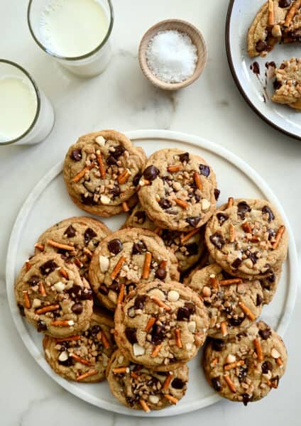 A top-down view of a white plate loaded with Kitchen Sink Cookies next to two glasses of milk and a small dish containing sea salt