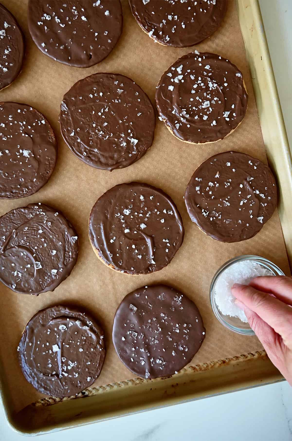 Sprinkling flaky sea salt atop chocolate-dipped cookies on a parchment paper-lined baking sheet.