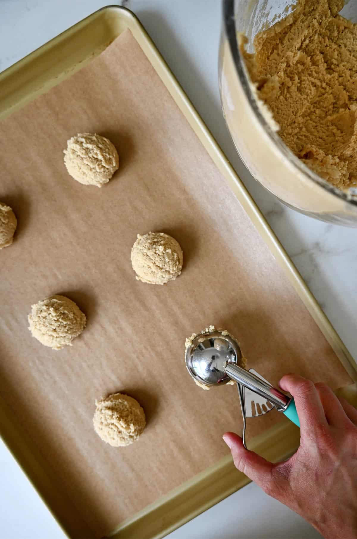 Using a cookie scoop to scoop chipless cookie dough balls onto a parchment paper-lined baking sheet.