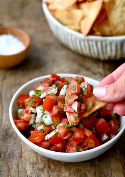 A hand holding a tortilla chip scoops up homemade pico de gallo from a small white bowl.